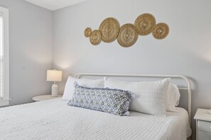 A bedroom with a bed, white bedding, a patterned pillow, a bedside lamp, and wall art. - Beach Bungalow - Walk to the Beach/Pool steps away (St. Augustine)