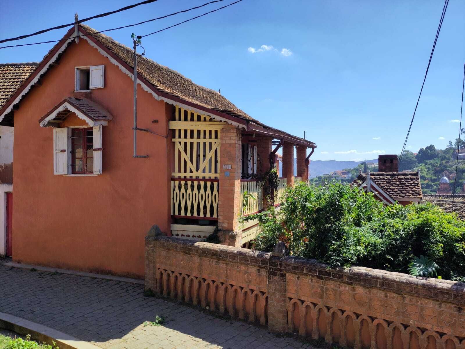 Ein zweistöckiges Haus mit roter Fassade, einem Balkon mit Holzgeländer und einer Ziegelwand davor.