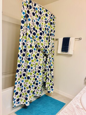 Um banheiro com cortina de chuveiro estampada com bolinhas, tapete de banho azul e toalha branca pendurada em barra de metal.