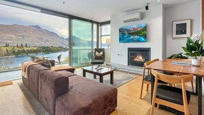 A modern living room with a large window, a sofa, a fireplace, and a dining area. - Iconic Lake View Apt | Ski Lockers (Queenstown)