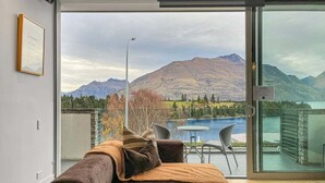 A modern living room with a sofa, a coffee table, and a view of mountains and a lake through large glass doors. - Iconic Lake View Apt | Ski Lockers (Queenstown)