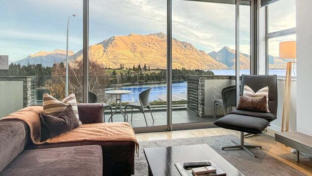 A modern living room with a large sliding glass door leading to a balcony with a view of mountains and a lake. - Iconic Lake View Apt | Ski Lockers (Queenstown)