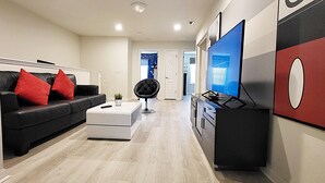 Una sala de estar moderna con un sofá de cuero negro, cojines rojos, un reposapiés blanco, una silla negra, un televisor de pantalla plana y una zona de cocina con un mueble negro y una pared de acento roja.