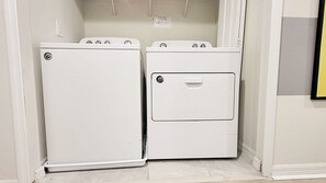 Een wasserette met twee gestapelde wasmachines en drogers, een betegelde vloer en een witte muur met een klein bordje boven de drogers.