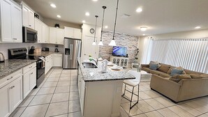 Una cocina moderna con armarios blancos, un refrigerador de acero inoxidable y una encimera de granito. Hay un rincón de comedor con un sofá marrón y un televisor montado en una pared de piedra.