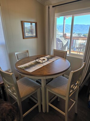 A dining area with a round table, four chairs, a framed picture on the wall, and a view of a deck and mountains through a window. - Panoramic Water Views - Pet Friendly &  Fisherman's Specials (Lakeport)