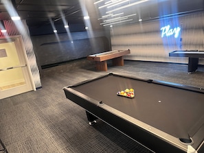 Ein Billardtisch mit Kugeln, eine hölzerne Bank und ein leuchtendes 'Play'-Schild in einem Raum mit Teppichboden und einer Wand mit vertikalen Linien.