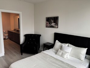 Ein Schlafzimmer mit Bett, Sessel, Nachttisch, Kommode und einem durchgehenden Tür sichtbaren Badezimmer.