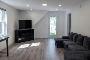 A modern living room with a flat-screen TV on a black entertainment unit, a grey sectional sofa, and a wooden floor.