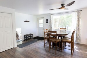 A dining area with a round table and six chairs, a ceiling fan, a door, and a window with curtains.