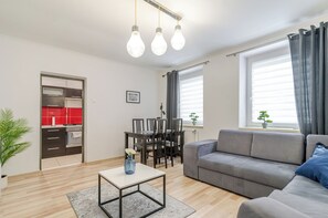 Ein modernes Wohnzimmer mit grauem Sofa, einem Esstisch mit Stühlen, einer Küche mit roten Akzenten und einem Fenster mit Vorhängen.