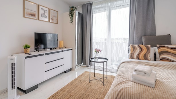 Habitación de hotel moderna con televisión de pantalla plana, una mesita con un florero, una cama con almohadones a rayas y un balcón con cortinas.