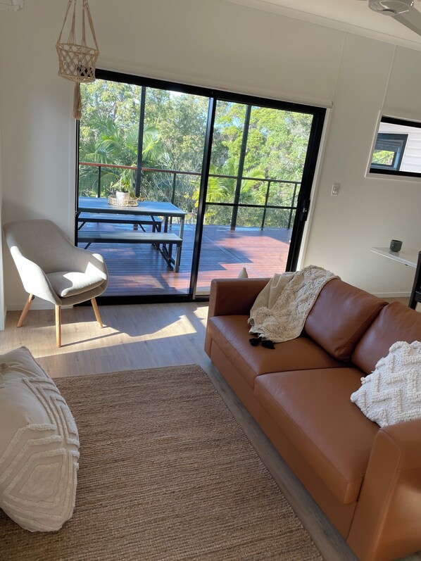 Uma sala de estar moderna com um sofá de couro marrom, uma poltrona branca, uma mesa de centro de madeira e uma grande porta de vidro deslizante que dá para um deck externo.