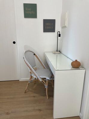 Quarto com escrivaninha branca, cadeira preta e branca, luminária na parede e duas obras de arte motivacionais.