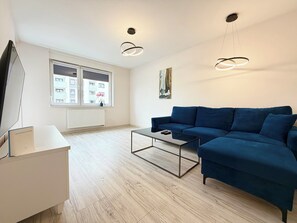 Ein modernes Wohnzimmer mit einem blauen Sofa, einem Glastisch, einer weißen Kommode und einem großen Fenster mit Blick auf Gebäude.