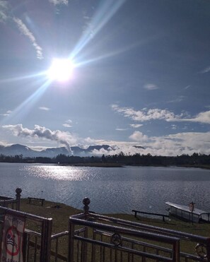 A lake with a metal railing in the foreground, a clear sky, and mountains in the background. - Cozzy Dira Cottage Pangalengan (Jawa Barat)