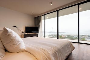 A modern hotel room with a large bed, a flat-screen TV, and a city view through floor-to-ceiling windows.