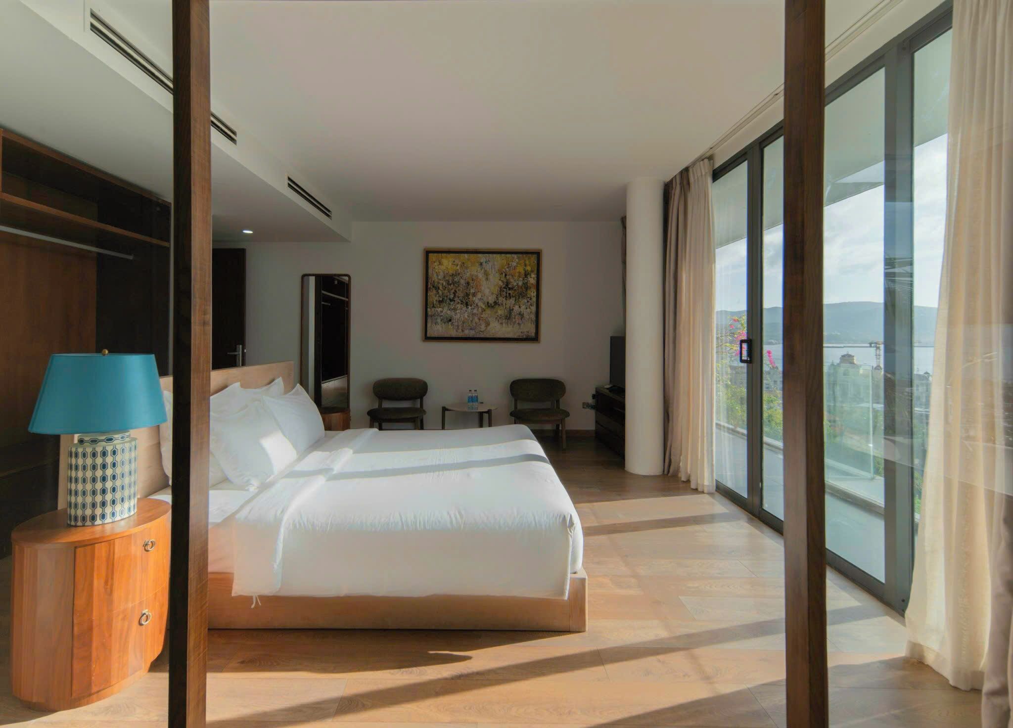 A hotel room with a large bed, a desk, a chair, a small table, a lamp, and a painting.