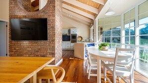 Una cucina con parete in mattoni, TV a schermo piatto, pavimento in legno e zona pranzo con sedie e tavolo bianchi.