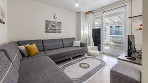 A modern living room with a grey sectional sofa, a white armchair, a flat-screen TV, and a large sliding glass door leading to an outdoor area. - Blue Bay Sea View Apartment With Free Parking (Himarë)