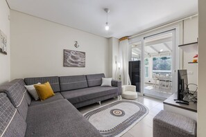 A modern living room with a grey sectional sofa, a white armchair, a flat-screen TV, and a large sliding glass door leading to an outdoor area. - Blue Bay Sea View Apartment With Free Parking (Himarë)