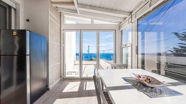 A modern kitchen with a stainless steel refrigerator, a dining table with a placemat, and a view of the ocean through large sliding glass doors. - Blue Bay Sea View Apartment With Free Parking (Himarë)