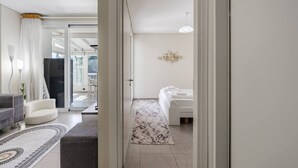A modern hotel room with a bed, a sitting area with a sofa and armchair, a hallway leading to a balcony, and a chandelier. - Blue Bay Sea View Apartment With Free Parking (Himarë)