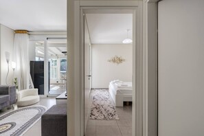 A modern hotel room with a bed, a sitting area with a sofa and armchair, a hallway leading to a balcony, and a chandelier. - Blue Bay Sea View Apartment With Free Parking (Himarë)