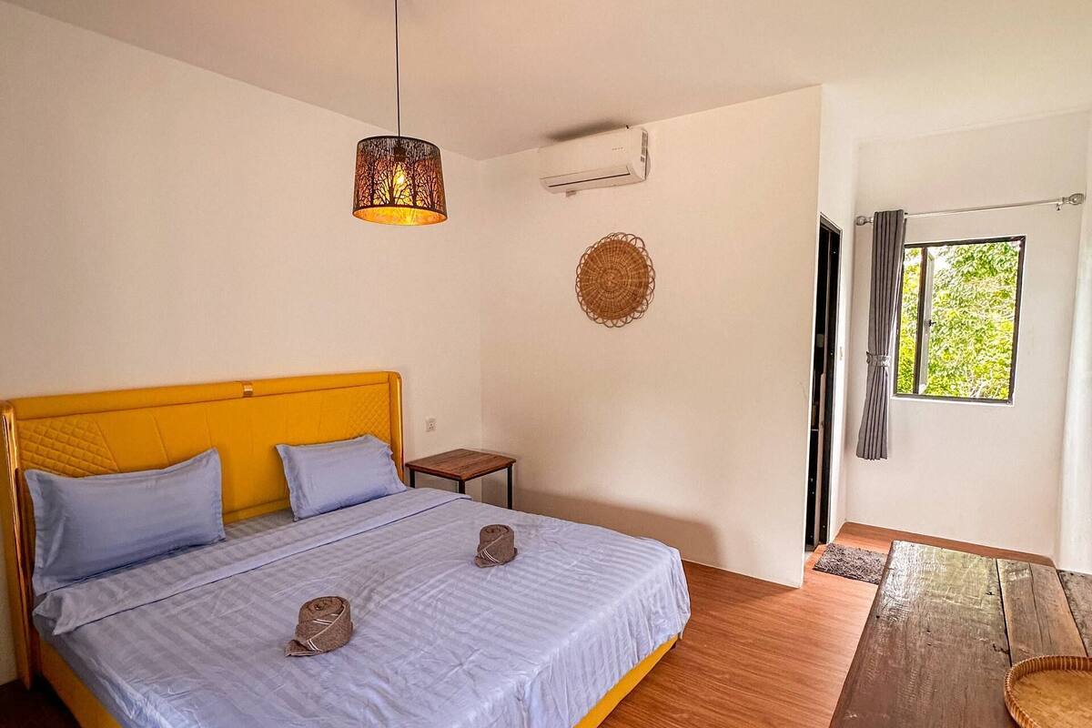 A room with a bed, a small table, a hanging light fixture, a woven wall decoration, and a window with curtains.