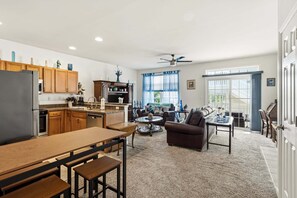 A modern kitchen with wooden cabinets, a dining area with a wooden table and benches, a living room with a sofa and coffee table, and a large window with blue curtains. - Relaxing First floor Condo with Outdoor Shower & Private Deck (Wildwood)
