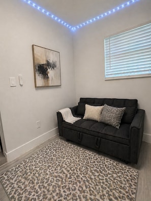 Une pièce avec un canapé noir, un tapis à motifs léopard, un tableau accroché au mur et une fenêtre avec des stores.