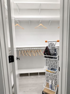 Un dressing accessible sans porte, avec un banc, des cintres en bois, un porte-chaussures et un chariot de nettoyage.