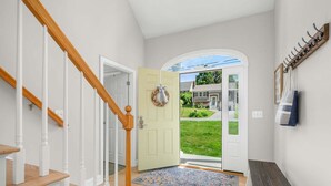 Un pasillo con una escalera de madera, una puerta verde claro, un felpudo y un banco.