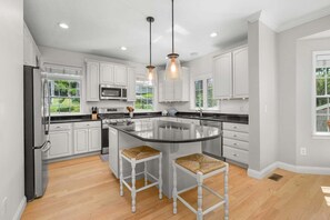 A modern kitchen with white cabinets, a large island, two bar stools, and pendant lighting. - Town Neck Beach Oasis (Sandwich)