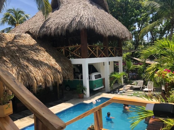 A pool area with a thatched-roof structure, a white building, and a green vehicle.