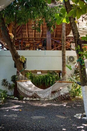 A hammock under a tree, a building with a thatched roof, and a white wall.