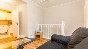 Ein modernes Wohnzimmer mit einem Glastisch, einem schwarzen Ledersofa und einem durchgehenden Gang sichtbarer Küchenbereich.