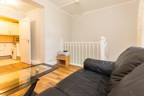 Ein modernes Wohnzimmer mit einem Glastisch, einem schwarzen Ledersofa und einem durchgehenden Gang sichtbarer Küchenbereich.