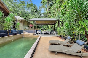 Ein Pool mit Liegestühlen und einer überdachten Sitzgelegenheit, umgeben von üppiger Vegetation.