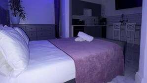 Une chambre à coucher moderne avec un lit, une table de chevet, une plante en pot et une vue sur un coin cuisine.