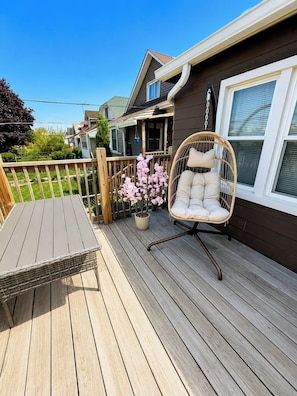 A wooden deck with a wicker chair and a table, a potted plant, and a house in the background.
