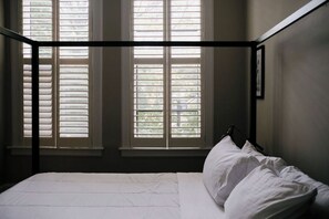 Ein Schlafzimmer mit Himmelbett, zwei Fenstern mit Jalousien und einem gerahmten Bild an der Wand.