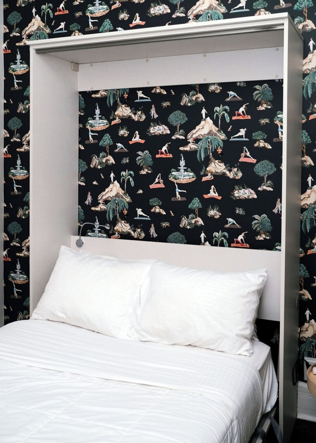 Quarto com cama, almofadas brancas e parede com fundo preto, exibindo ilustrações de pessoas e natureza.