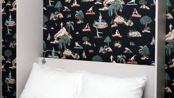 Quarto com cama, almofadas brancas e parede com fundo preto, exibindo ilustrações de pessoas e natureza.
