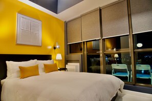 Een hotelkamer met een groot bed, uitzicht op de stad door middel van vloerhoogte ramen, een wandlamp en een raambekleding.