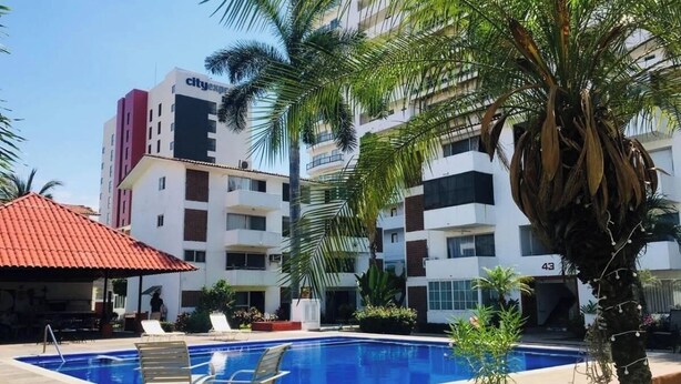 A swimming pool with lounge chairs, palm trees, and a building with balconies. - Apartment with pool near the beach in PV (Puerto Vallarta)