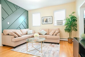 Ein Wohnzimmer mit Ecksofa, Glastisch, Topfpflanze und Kunstwerken an den Wänden.