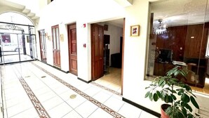Un pasillo de hotel con suelo de baldosas, puertas de madera, una planta en maceta y un ventanal grande.