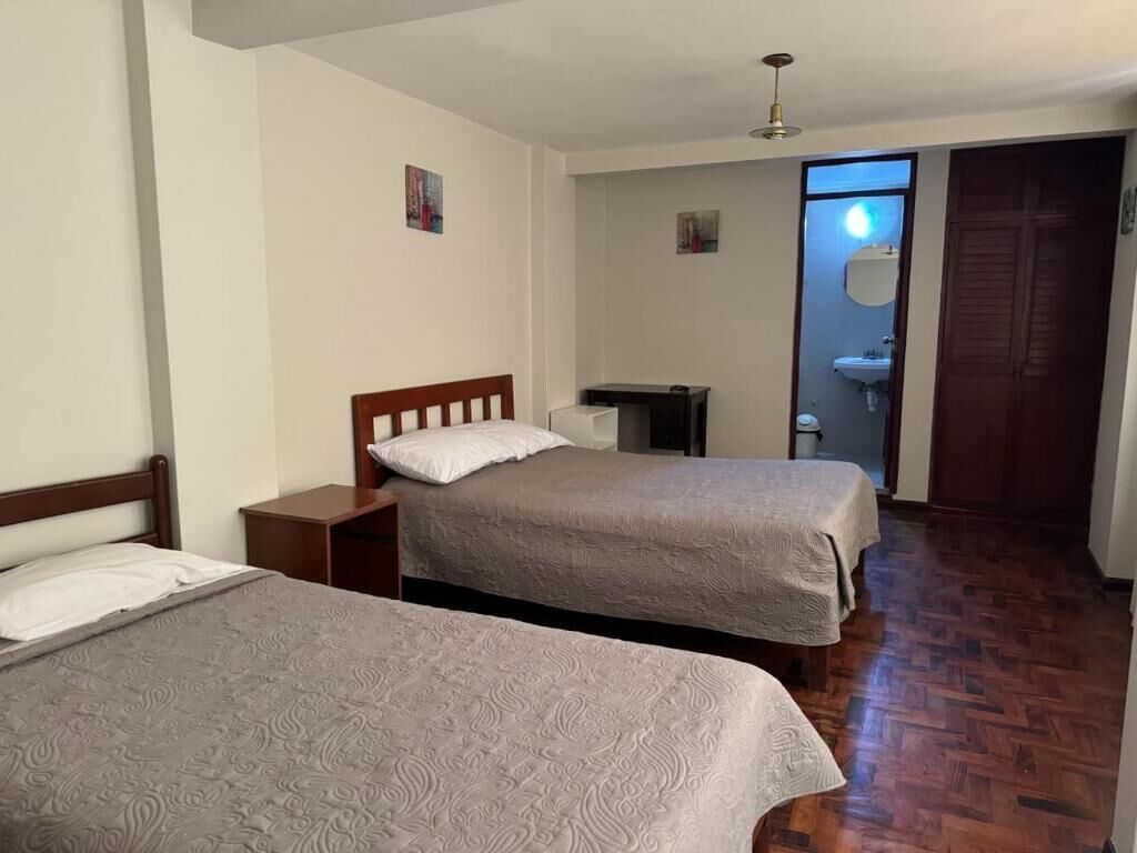 A room with two beds, a wooden floor, a desk, a chair, and a door leading to a bathroom.