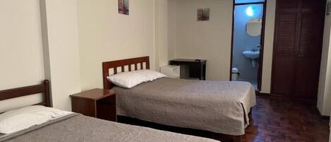 A room with two beds, a wooden floor, a desk, a chair, and a door leading to a bathroom.